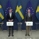 İsveç Hükümeti basın toplantısında "vandel" dürüst yaşam kriterlerini açıklayan dört erkek yetkili podyumda duruyor, İsveç ve AB bayrakları arka planda. İsveç Hükümet Ofisi (Regeringskansliet) logosunu taşıyan dört şeffaf podyumun arkasında, resmi kıyafetli dört erkek yetkili duruyor. Soldan ikinci olan kişi (sarı kravatlı) konuşuyor. Arka planda dönüşümlü olarak sıralanmış İsveç bayrakları ve Avrupa Birliği bayrakları yer alıyor. Fotoğraf, İsveç'teki resmi bir hükümet basın açıklamasını yansıtıyor.