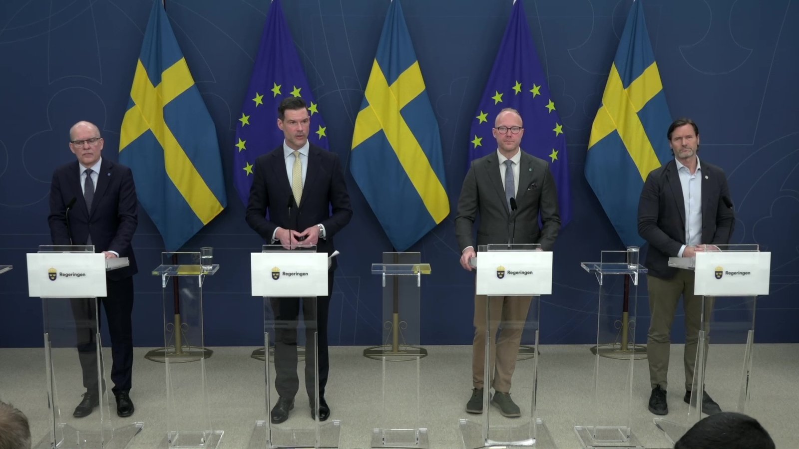 İsveç Hükümeti basın toplantısında "vandel" dürüst yaşam kriterlerini açıklayan dört erkek yetkili podyumda duruyor, İsveç ve AB bayrakları arka planda. İsveç Hükümet Ofisi (Regeringskansliet) logosunu taşıyan dört şeffaf podyumun arkasında, resmi kıyafetli dört erkek yetkili duruyor. Soldan ikinci olan kişi (sarı kravatlı) konuşuyor. Arka planda dönüşümlü olarak sıralanmış İsveç bayrakları ve Avrupa Birliği bayrakları yer alıyor. Fotoğraf, İsveç'teki resmi bir hükümet basın açıklamasını yansıtıyor.